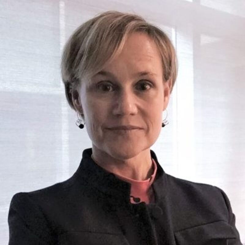 The image shows a woman with short blonde hair, wearing a black jacket over a pink shirt. She has a neutral expression and is looking directly at the camera. The background is a blurred white, suggesting an indoor setting. She appears to be in her late 40s or early 50s.